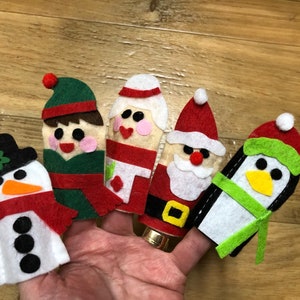 May include: Five felt finger puppets depicting Christmas characters. The puppets are a snowman, an elf, a woman wearing a red dress, Santa Claus, and a penguin.