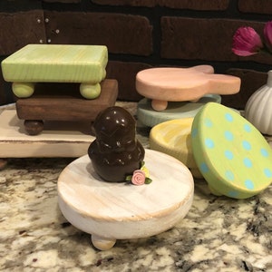 May include: A collection of six wooden coasters with various pastel colors and designs, including a bunny, polka dots, and swirls. The coasters are displayed on a granite countertop with a chocolate bunny and a small bouquet of pink flowers.