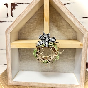 May include: A small, rustic wreath with a black and white checkered bow hanging in a wooden house-shaped shelf.