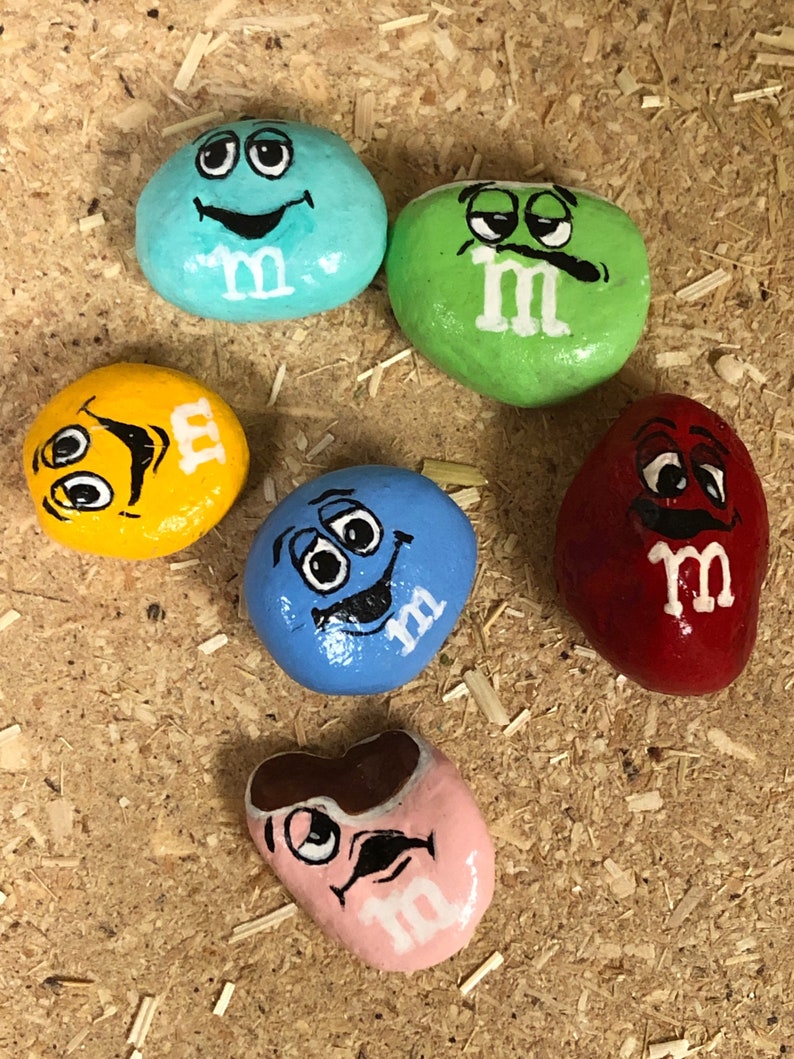 May include: Six painted rocks with different colors and faces, each with the letter "M" painted on them. The rocks are painted to look like M&M's candies.
