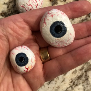 May include: Three painted rock eyeballs with red veins and blue irises. The eyeballs are different sizes and are held in a hand.