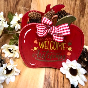 May include: Red wooden apple shaped sign with a white and red gingham bow and pine cones. The sign says "So Long Summer, Welcome Autumn".