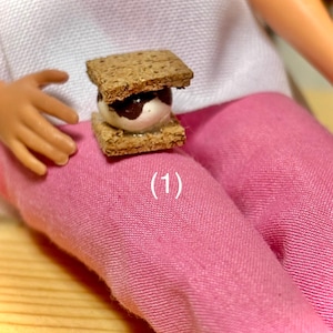 May include: A miniature s'more, featuring two graham cracker squares with a marshmallow and chocolate filling. The s'more is resting on a pink surface. The number (1) is visible on the pink surface.