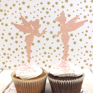 Puede incluir: Dos toppers de cupcake de hada de purpurina rosa sobre cupcakes con chispas rosas. El fondo es un papel blanco con estrellas doradas.