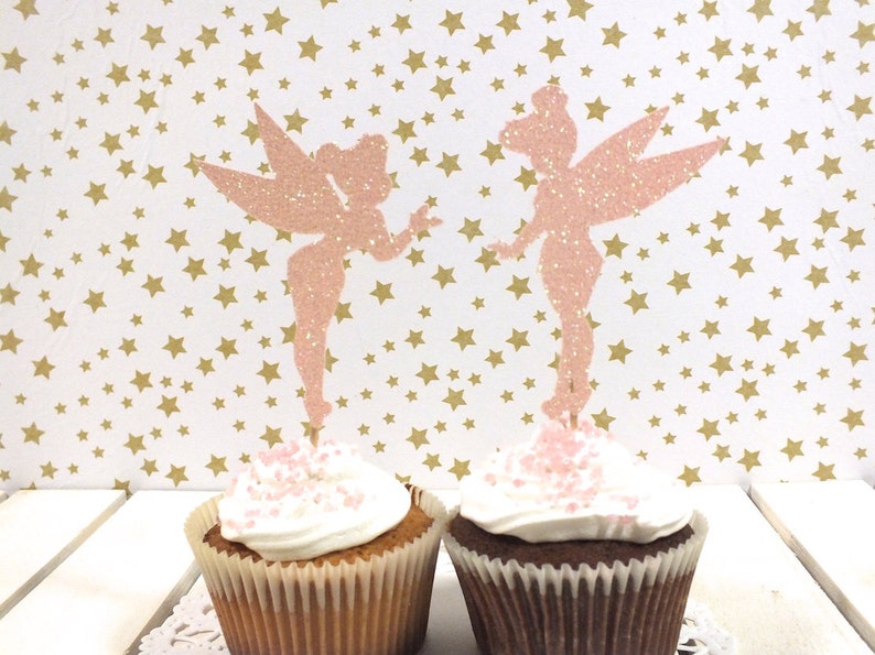 Puede incluir: Dos toppers de cupcake de hada de purpurina rosa sobre cupcakes con chispas rosas. El fondo es un papel blanco con estrellas doradas.
