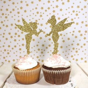Puede incluir: Dos cupcakes con adornos de campanilla de oro brillante. Los cupcakes est&aacute;n cubiertos con glaseado blanco y chispas rosas.
