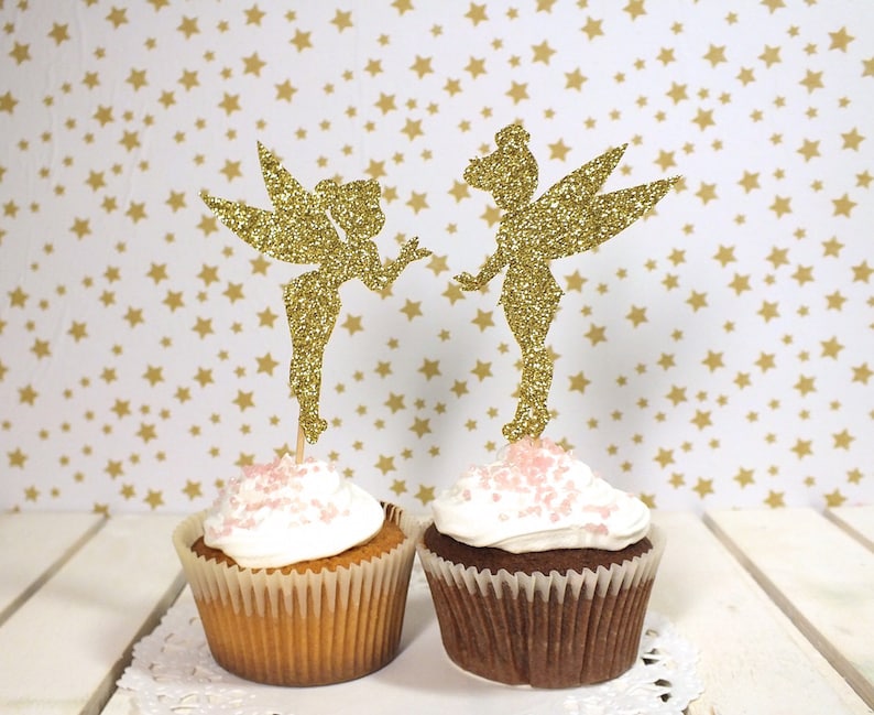 Puede incluir: Dos cupcakes con adornos de campanilla de oro brillante. Los cupcakes est&aacute;n cubiertos con glaseado blanco y chispas rosas.
