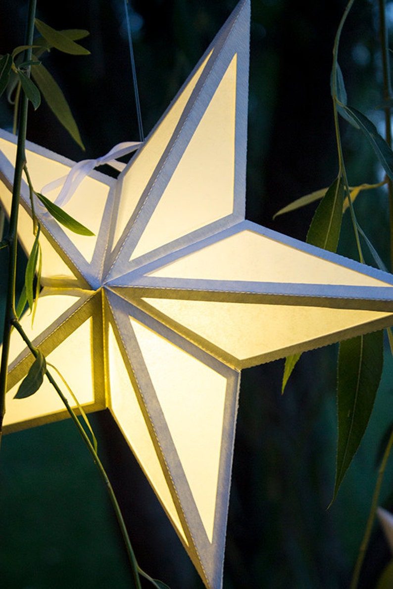 Op de afbeelding: Een witte papieren sterlantaarn met een licht erin, hangend aan een tak met groene bladeren.
