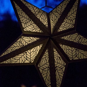 Puede incluir: Una linterna de papel en forma de estrella negra con un patrón de hojas blancas. La linterna está encendida y brilla, proyectando una luz cálida.