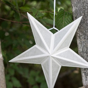 Op de afbeelding: Een witte papieren stervormige lantaarn die aan een wit touwtje hangt. De ster is gemaakt van papier en heeft een 3D-ontwerp. De ster hangt voor een houten ladder en groen gebladerte.