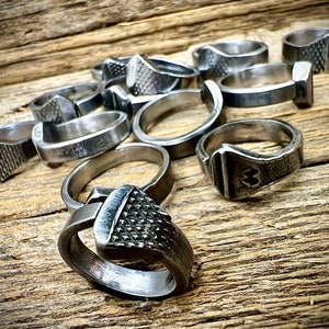 May include: A group of silver metal rings, each with a unique design, are arranged on a wooden surface. The rings are made from recycled materials and are handcrafted.