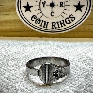 May include: A silver ring with a unique design featuring a textured, rectangular shape with two small circles. The ring is on a white, textured surface.