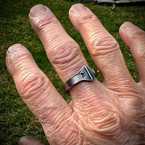 May include: A silver ring with a unique design featuring a small, raised, geometric shape. The ring is worn on the middle finger of a person's hand.