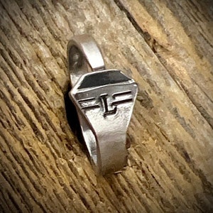 May include: A silver ring with a black enamel inlay and a stylized letter "F" with wings. The ring is on a wooden surface.