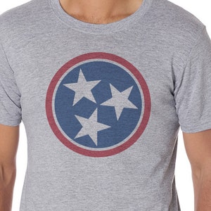 May include: A gray t-shirt with a blue and red circle featuring three white stars. The circle is a stylized version of the Tennessee state flag.