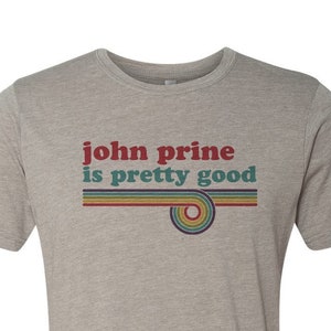 Puede incluir: Una camiseta gris jaspeada con el texto "john prine is pretty good" en una fuente retro con rayas arcoíris.