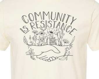 May include: A cream colored t-shirt with a black graphic print that reads "Community is Resistance". The graphic features a handshake with flowers growing around it.