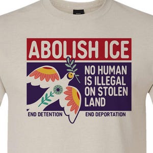 Puede incluir: Una camiseta de color beige claro con un diseño gráfico. El diseño presenta las palabras "ABOLISH ICE" en rojo, una paloma con una rama de olivo y el texto "NO HUMAN IS ILLEGAL ON STOLEN LAND". La camiseta también dice "END DETENTION" y "END DEPORTATION".