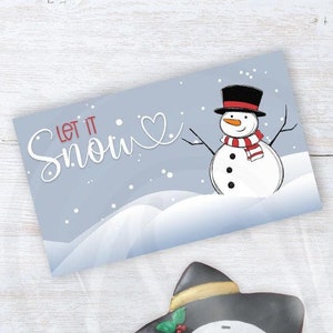 May include: A star-shaped snowman cookie with a black hat, red scarf, and carrot nose, next to a card that says "Let it Snow" with a snowman illustration. The cookie is in a clear bag. The card is light blue.