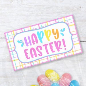 Puede incluir: Topper para bolsa de regalo rosa y amarillo "Feliz Pascua" con un patrón de cuadros. El topper está diseñado para doblarse sobre una bolsa de regalo de 10 cm o 16,5 cm. La imagen también muestra una bolsa de plástico transparente llena de huevos de caramelo de colores.