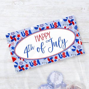 Peut inclure: Un sac en plastique transparent rempli de bonbons, avec une carte décorative sur le dessus. La carte présente un motif bleu et rouge avec le texte "HAPPY 4th of July" et "USA". Le sac contient des bonbons Reese's.