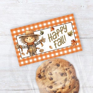 May include: A chocolate chip cookie in a clear bag next to a rectangular card with an orange and white checkered pattern. The card features a scarecrow illustration and the text "Happy Fall!"
