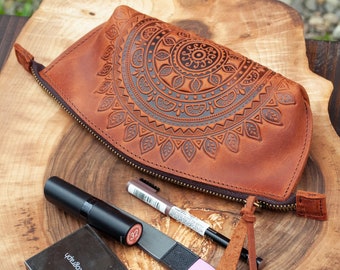 May include: A brown leather cosmetic bag with a detailed mandala design, a black mascara tube, a compact, a nail file, and a brown pen are displayed on a wooden surface. The bag has a zipper closure and a leather pull.