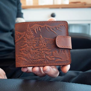 Puede incluir: Cartera de cuero marrón con un diseño de montaña y pinos. La cartera tiene un cierre de botón a presión y está sujeta en la mano de una persona. La cartera es de color marrón intenso.