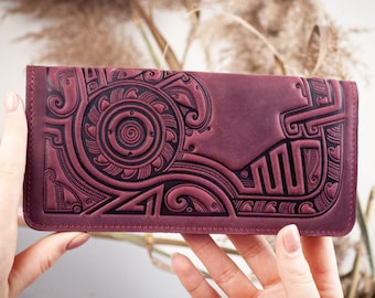 May include: A burgundy leather wallet with an intricate, embossed design. The design features geometric and swirling patterns, creating a detailed and textured surface. The wallet is rectangular and appears to be closed, showcasing the craftsmanship and artistry.