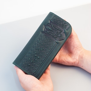 Puede incluir: Cartera de cuero verde oscuro con un diseño geométrico y estilizado en relieve. La cartera rectangular presenta un patrón detallado en la parte delantera. Las costuras son visibles a lo largo de los bordes. La cartera se sostiene con dos manos.