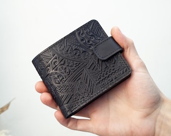 Peut inclure: Portefeuille en cuir noir avec un motif géométrique en relief. Le portefeuille a une fermeture à bouton-pression et est tenu dans une main. Le design présente diverses formes et lignes, créant un aspect texturé. C'est un portefeuille à deux volets.