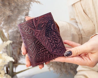 Peut inclure: Un portefeuille en cuir bordeaux avec des motifs géométriques et abstraits en relief. Le portefeuille présente des coutures rouges et un design détaillé, mettant en valeur une esthétique unique et artisanale. Le portefeuille est tenu dans les mains.