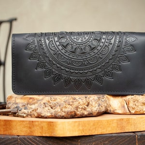Könnte beinhalten: Schwarze Ledergeldbörse mit geprägtem Mandala-Design. Die rechteckige Geldbörse weist ein detailliertes, symmetrisches Muster auf. Sie wird auf einer Holzoberfläche präsentiert, um ihre Handwerkskunst und ihren Stil hervorzuheben.