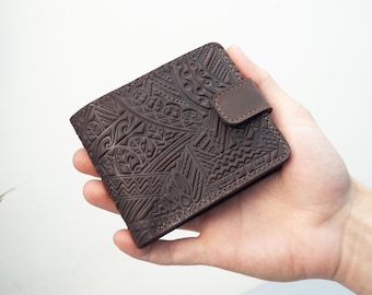 Peut inclure: Un portefeuille en cuir marron avec un motif géométrique en relief complexe. Le portefeuille a une fermeture à bouton-pression et est tenu dans une main sur fond blanc. Le design présente diverses formes et lignes.