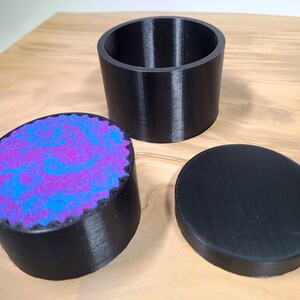K&ouml;nnte beinhalten: Dreiteiliges schwarzes Beh&auml;lterset. Das Set enth&auml;lt einen zylindrischen offenen Beh&auml;lter, einen Deckel und einen kleineren Beh&auml;lter mit einem wirbelnden lila und blauen Design. Die Beh&auml;lter bestehen aus einem strukturierten Material und liegen auf einer Holzoberfl&auml;che.