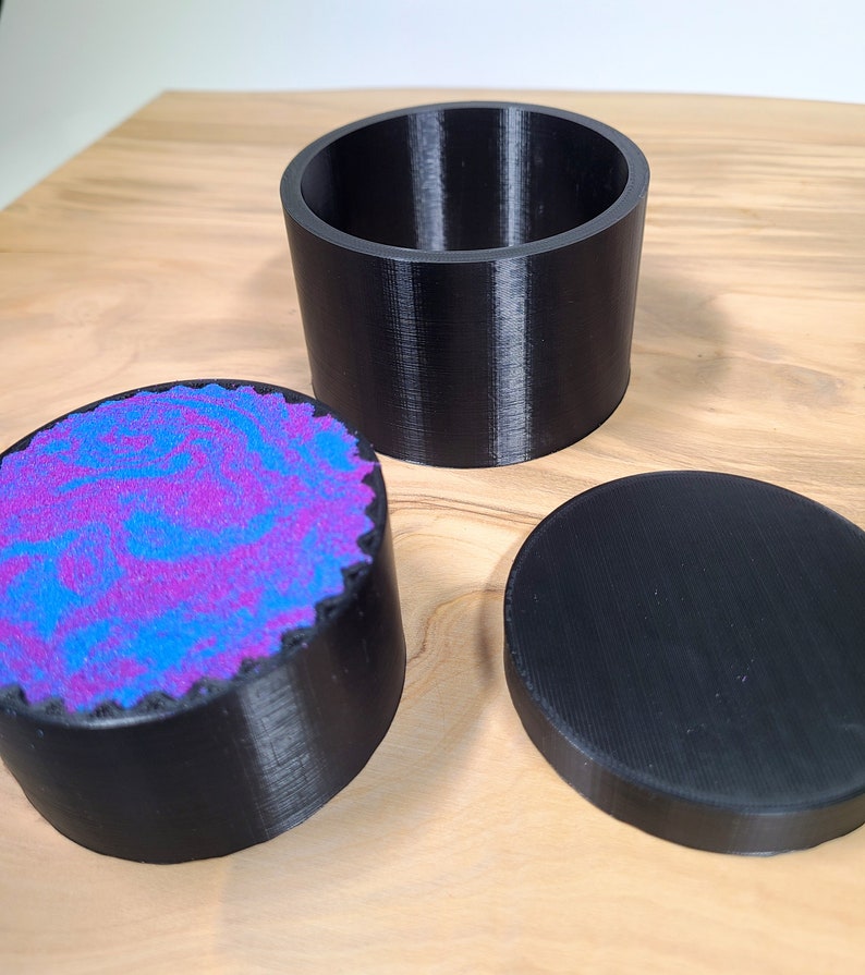 K&ouml;nnte beinhalten: Dreiteiliges schwarzes Beh&auml;lterset. Das Set enth&auml;lt einen zylindrischen offenen Beh&auml;lter, einen Deckel und einen kleineren Beh&auml;lter mit einem wirbelnden lila und blauen Design. Die Beh&auml;lter bestehen aus einem strukturierten Material und liegen auf einer Holzoberfl&auml;che.