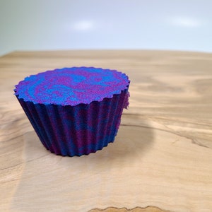 K&ouml;nnte beinhalten: Ein cupcake-f&ouml;rmiger Badeschaum mit gewelltem Rand. Der Badeschaum hat einen Wirbel aus blauen und violetten Farben mit einer dunkelblauen Basis. Das Objekt befindet sich auf einer Holzoberfl&auml;che.