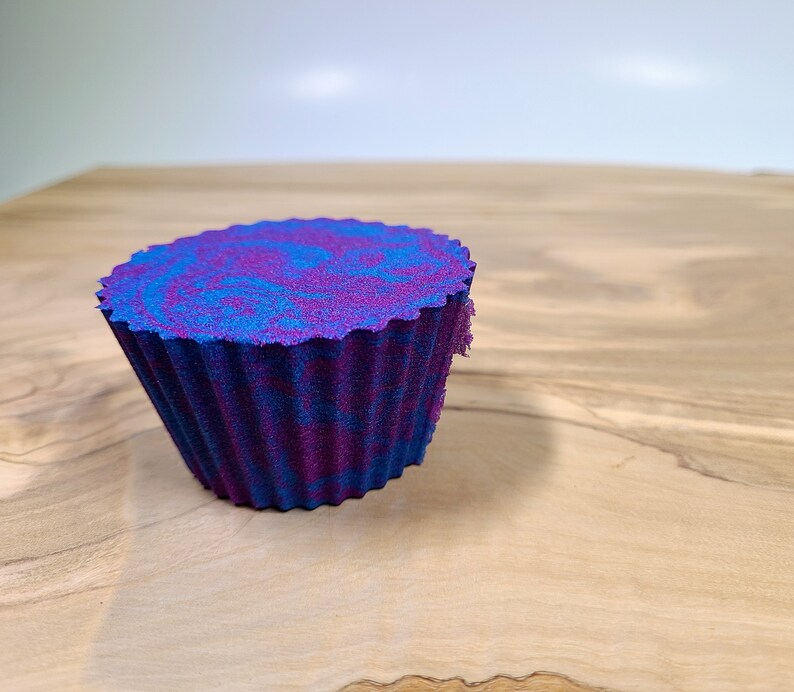 K&ouml;nnte beinhalten: Ein cupcake-f&ouml;rmiger Badeschaum mit gewelltem Rand. Der Badeschaum hat einen Wirbel aus blauen und violetten Farben mit einer dunkelblauen Basis. Das Objekt befindet sich auf einer Holzoberfl&auml;che.