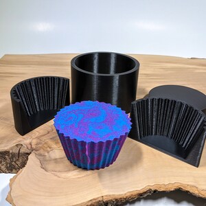 K&ouml;nnte beinhalten: Eine Sammlung von 3D-gedruckten Formen und ein farbenfroher, gewirbelter Cupcake. Die Formen sind schwarz und umfassen eine zylindrische Form, zwei Halbkreisformen und eine kreisf&ouml;rmige Oberseite. Der Cupcake hat ein blaues und violettes Wirbeldesign.