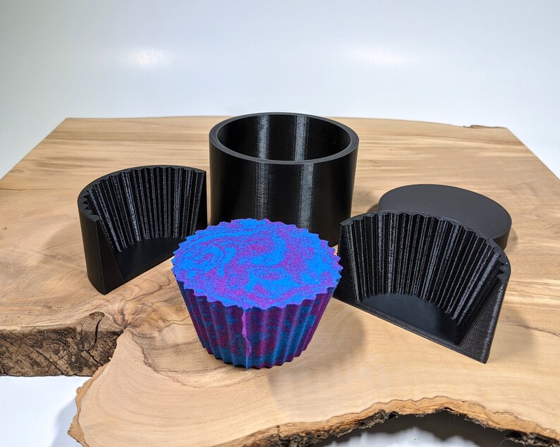 K&ouml;nnte beinhalten: Eine Sammlung von 3D-gedruckten Formen und ein farbenfroher, gewirbelter Cupcake. Die Formen sind schwarz und umfassen eine zylindrische Form, zwei Halbkreisformen und eine kreisf&ouml;rmige Oberseite. Der Cupcake hat ein blaues und violettes Wirbeldesign.