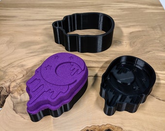 May include: Black plastic skull-shaped cookie cutter with a purple bath bomb in the shape of a skull with a crescent moon and stars. The bath bomb is next to the cutter. The items are on a wooden surface.