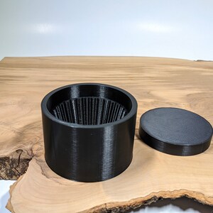 K&ouml;nnte beinhalten: Ein schwarzer, zylindrischer Beh&auml;lter mit geriffeltem Innenraum und einem separaten, flachen Deckel. Der Beh&auml;lter und der Deckel bestehen aus mattem, schwarzem Material. Die Artikel werden auf einer hellbraunen Holzoberfl&auml;che pr&auml;sentiert.