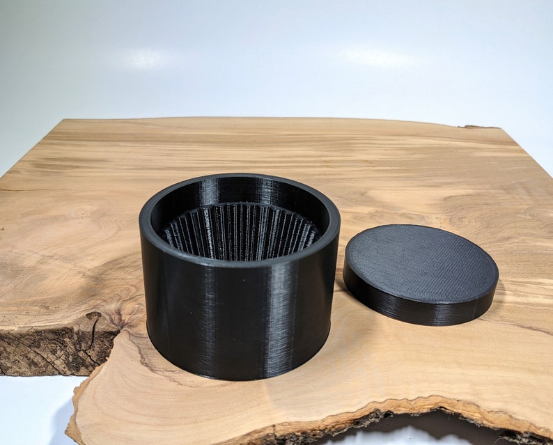 K&ouml;nnte beinhalten: Ein schwarzer, zylindrischer Beh&auml;lter mit geriffeltem Innenraum und einem separaten, flachen Deckel. Der Beh&auml;lter und der Deckel bestehen aus mattem, schwarzem Material. Die Artikel werden auf einer hellbraunen Holzoberfl&auml;che pr&auml;sentiert.