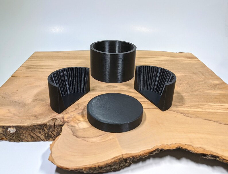 K&ouml;nnte beinhalten: Ein Satz von vier schwarzen 3D-gedruckten Objekten. Das Set beinhaltet einen zylindrischen Beh&auml;lter, eine flache kreisf&ouml;rmige Scheibe und zwei halbkreisf&ouml;rmige Teile mit vertikalen Rippen. Die Objekte sind auf einer Holzoberfl&auml;che angeordnet.