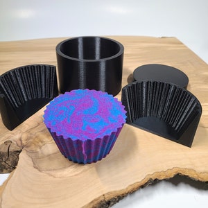 K&ouml;nnte beinhalten: Ein farbenfroher Badezusatz in Cupcake-Form mit blau-violetten Wirbeln liegt auf einem Holzbrett. Daneben befinden sich schwarze Kunststoffformen: ein Zylinder, zwei Halbkreisformen und ein kreisf&ouml;rmiger Deckel. Der Badezusatz hat einen strukturierten, ger&uuml;schten Rand.