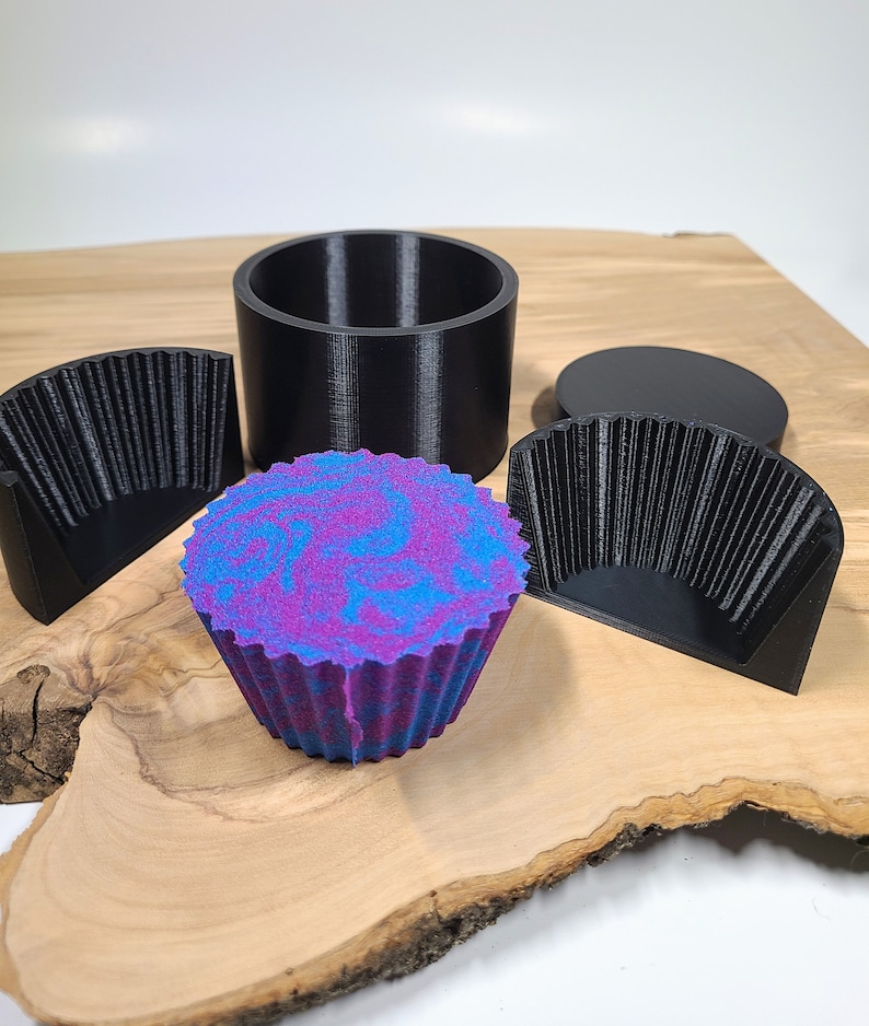 K&ouml;nnte beinhalten: Ein farbenfroher Badezusatz in Cupcake-Form mit blau-violetten Wirbeln liegt auf einem Holzbrett. Daneben befinden sich schwarze Kunststoffformen: ein Zylinder, zwei Halbkreisformen und ein kreisf&ouml;rmiger Deckel. Der Badezusatz hat einen strukturierten, ger&uuml;schten Rand.