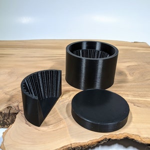 K&ouml;nnte beinhalten: Schwarze, 3D-gedruckte Grinder-Komponenten, darunter eine zylindrische Basis, ein Deckel und ein viertelkreisf&ouml;rmiges Teil. Die Grinder-Teile sind auf einer Holzoberfl&auml;che angeordnet.