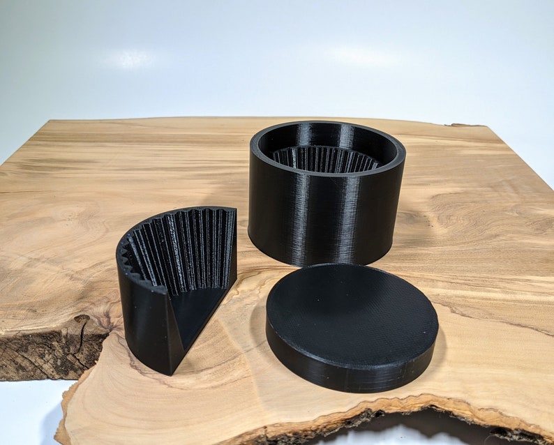 K&ouml;nnte beinhalten: Schwarze, 3D-gedruckte Grinder-Komponenten, darunter eine zylindrische Basis, ein Deckel und ein viertelkreisf&ouml;rmiges Teil. Die Grinder-Teile sind auf einer Holzoberfl&auml;che angeordnet.