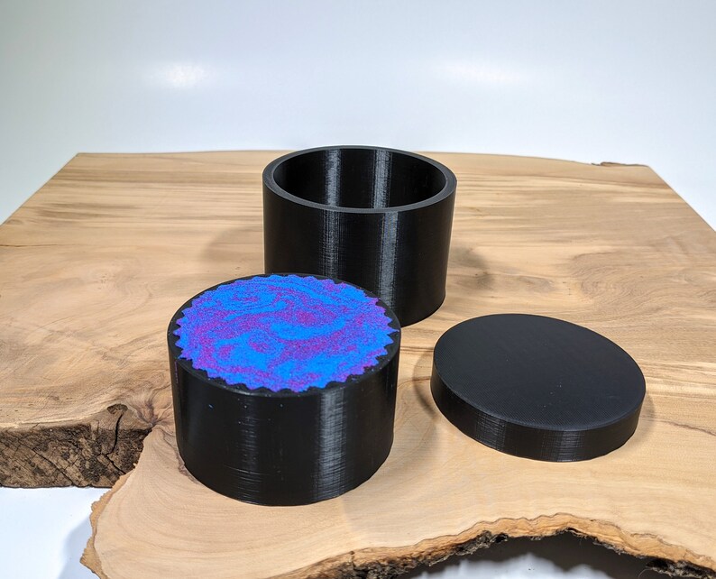 K&ouml;nnte beinhalten: Dreiteiliges schwarzes Beh&auml;lterset. Das Set beinhaltet einen zylindrischen Sockel mit blau-violett gesprenkelter Oberseite, einen passenden Deckel und ein separates zylindrisches Teil. Die Artikel werden auf einer Holzoberfl&auml;che pr&auml;sentiert.
