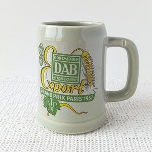 May include: A grey ceramic beer stein with a yellow and green logo that reads "Dortmunder DAB Aktienbrauerei Export Grand Prix Paris 1937".